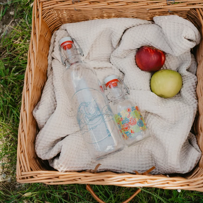 Zwei unterschiedlich bedruckte Glasflaschen liegen auf einer Decke in einem Picknickkorb neben einem grünen und einem roten Apfel