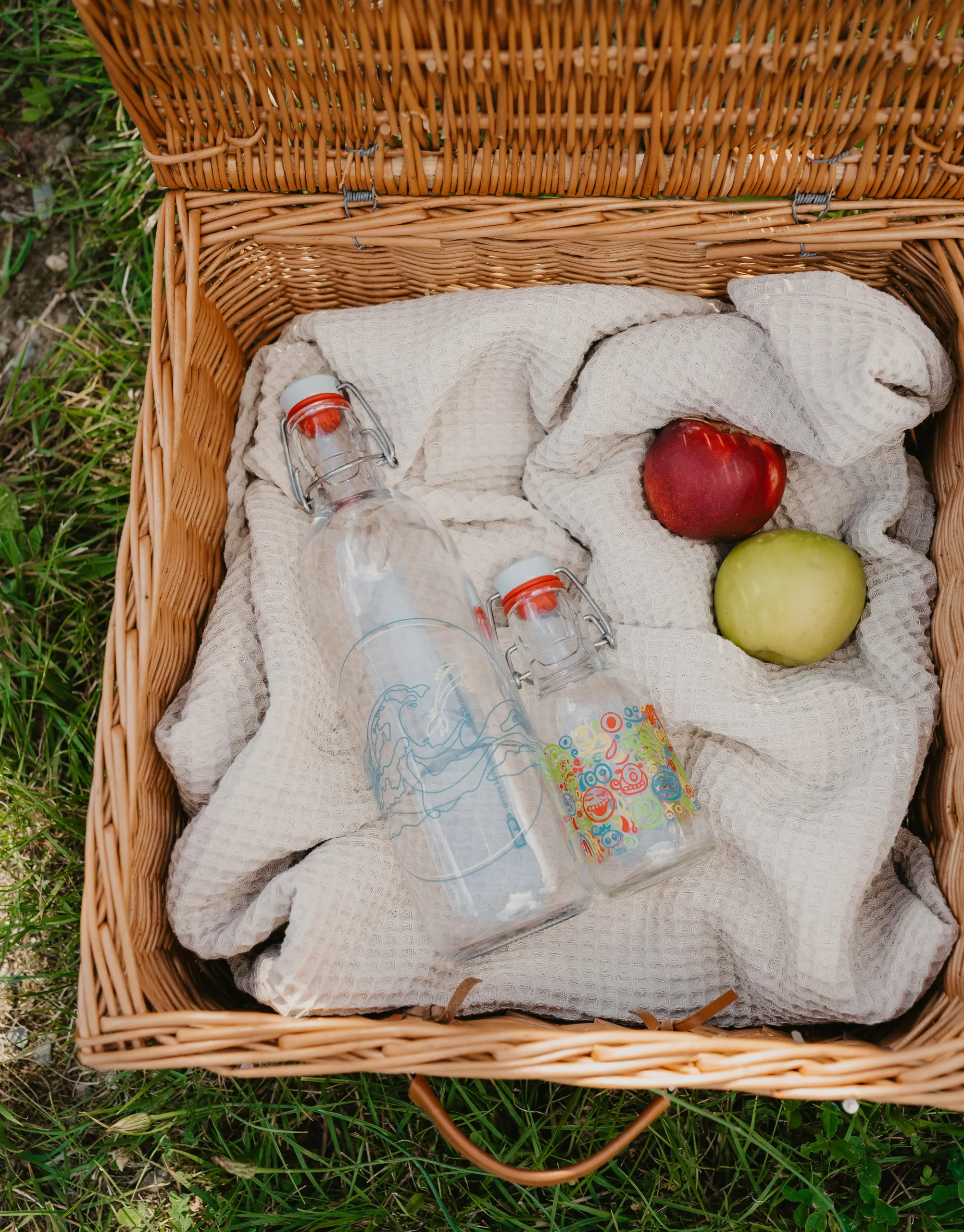 Zwei unterschiedlich bedruckte Glasflaschen liegen auf einer Decke in einem Picknickkorb neben einem grünen und einem roten Apfel