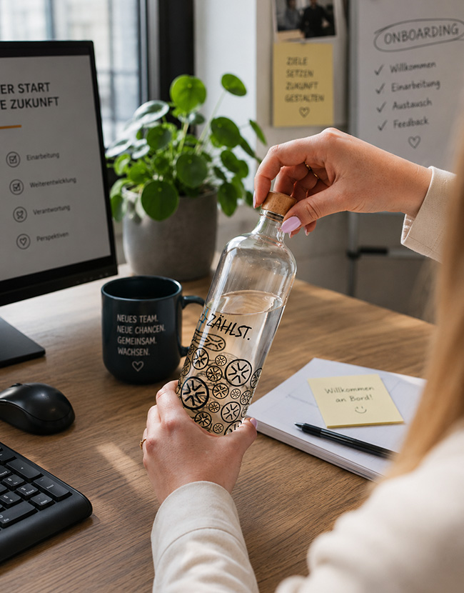 Büromitarbeiter schraubt individuell bedruckte Flasche an seinem Schreibtisch auf