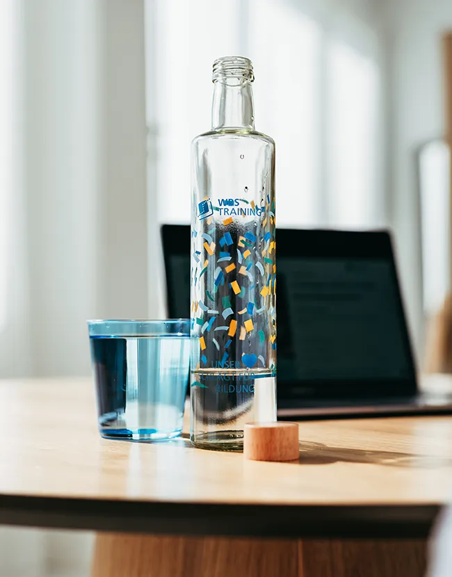 Im Siebdruck bedruckte Glasflasche auf einem Holztisch neben einem Laptop und einem gefüllten Wasserglas